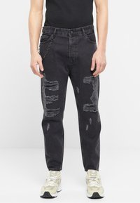 Jeans de mezclilla negra con detalles desgastados y un corte recto. Presentan una cadena en el lazo del cinturón frontal. Combinados con zapatillas de color claro.