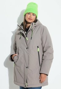 Giacca isolante grigio chiaro con cappuccio, dotata di tasche con zipper nere e dettagli in cordino verde brillante. Indossata con un berretto verde abbinato.