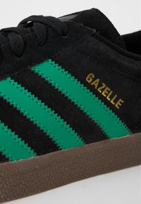 Crne suede tenisice s tri teksturirane zelene pruge, zlatnim natpisom "GAZELLE" i teksturiranom smeškom gumom.