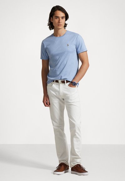 Gaiši zila kokvilnas T-kreklā ar apaļu kakla izgriezumu, baltas džinsus, brūnas ādas kurpes un brūnu jostu, modele stāv pret vieglu fonu.