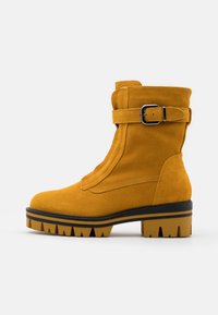 Botte chevillère en daim jaune avec une sangle à boucle, surface texturée et épais talon robuste avec un design en encoches. Détail de fermeture éclair sur le côté.
