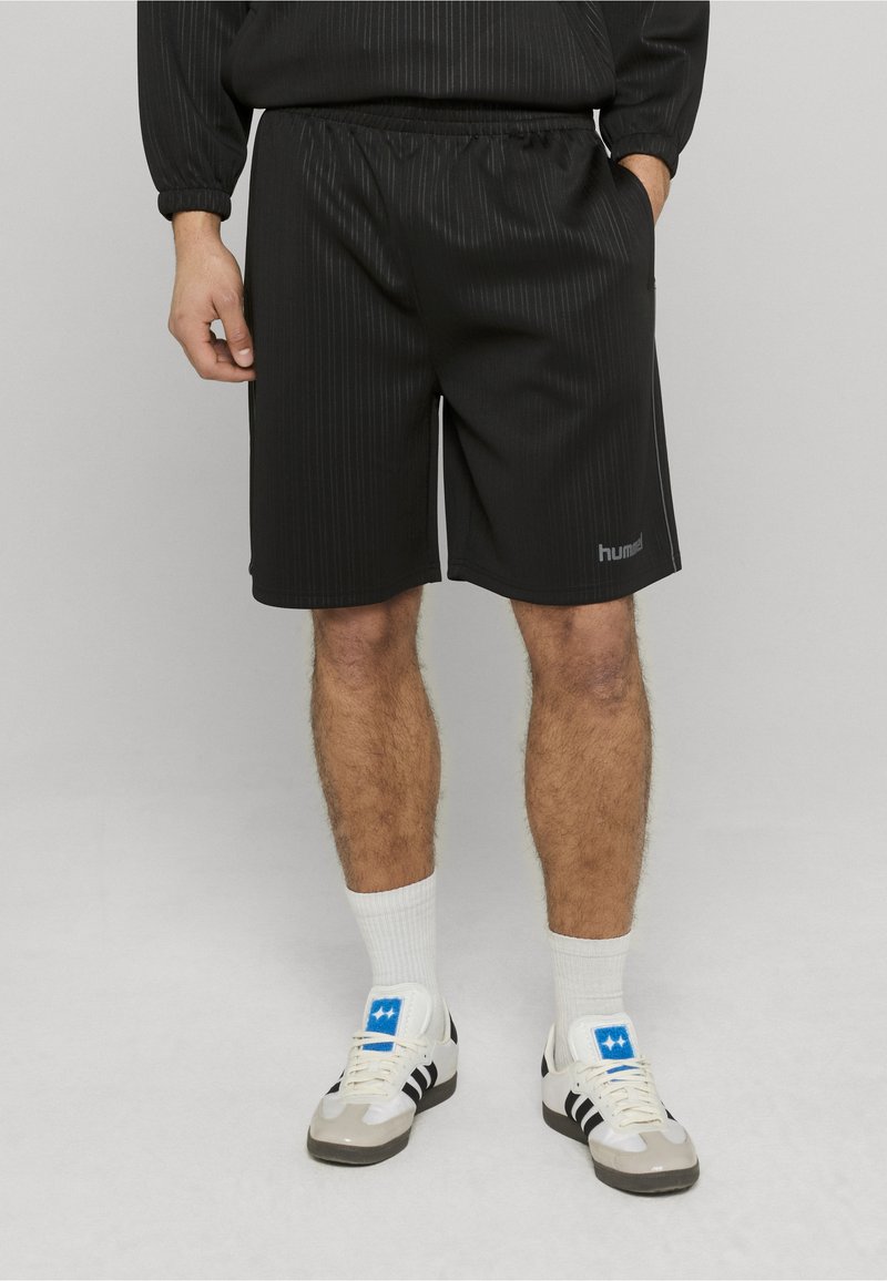 Shorts de sport noirs à texture rayée, taille élastique, poches latérales et petit logo. Portés avec des chaussettes blanches et des baskets.