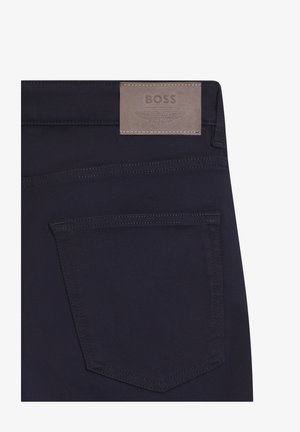Pantaloni in tessuto blu navy con tasca posteriore e una toppa rettangolare in pelle con l'etichetta "BOSS" e un logo alato in rilievo. Texture liscia e cuciture pulite.