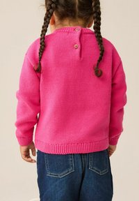 Bambina con due trecce codino, che indossa un vivace maglione rosa con bottoni e jeans blu, di spalle su uno sfondo neutro.