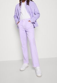 Fialová bavlněná košile spárovaná s odpovídajícími fialovými bavlněnými džínami. Pod ní bílý tričko a outfit doplňují bílé tenisky.