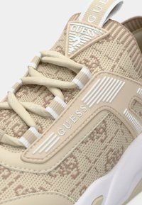 Baskets beiges avec une tige en maille texturée, des accents blancs et un patch de marque bien visible. Elles présentent un col rembourré et des lacets en croix.