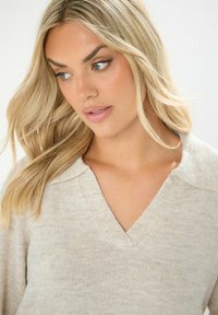 Hellbeige Pullover mit V-Ausschnitt und weitem Kragen, aus einem strukturierten Strickstoff gefertigt. Weicher, lässiger Stil mit einer lockeren Passform.