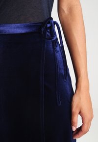 Gonna in velluto blu navy con chiusura laterale a legaccio. Presenta una texture liscia e una silhouette slim. Vista ravvicinata dei dettagli in vita.