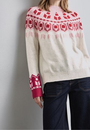 Femme portant un pull crème avec des motifs géométriques roses et rouges sur le plastron et les poignets, associé à un jean bleu foncé.