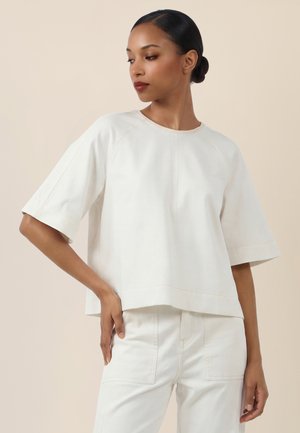 Frau mit dunklen Haaren in einem niedrigen Dutt, die ein übergroßes weißes Kurzarmoberteil und weiße Hose trägt, blickt nach links mit der Hand auf der Hüfte.