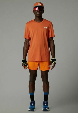 Camiseta deportiva naranja y pantalones cortos, combinados con una gorra y gafas de sol. El conjunto presenta detalles contrastantes en negro y neón, diseñado para uso activo.