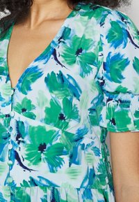 Blommig klänning i ljusblått med gröna och blå akvarellmönster. Har puffärmar, en djup V-ringning och knapppynt framtill.