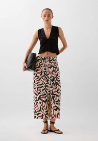 Schwarzes, verkürztes Oberteil mit V-Ausschnitt; gemusterte, weit geschnittene Hose in Creme, Schwarz und Pink; schwarze Tasche; flache Sandalen mit Knöchelriemen.