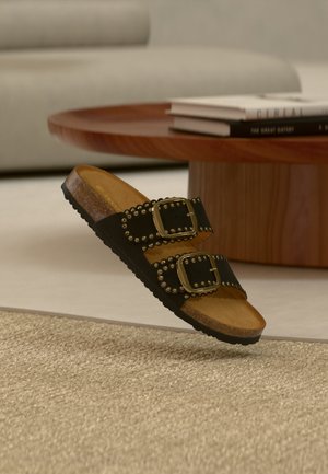 Μαύρο σανδάλι με σαγιονάρα με δύο λουριά με καρφιά και χρυσά αγκράφες που ακουμπούν σε μπεζ χαλί κοντά σε στρογγυλό ξύλινο τραπεζάκι καφέ με βιβλία.