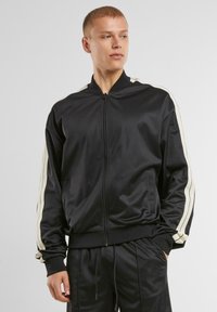 Veste zippée noire en matériau lisse, avec des rayures blanches le long des épaules et des manches, des poignets côtelés et un col montant.