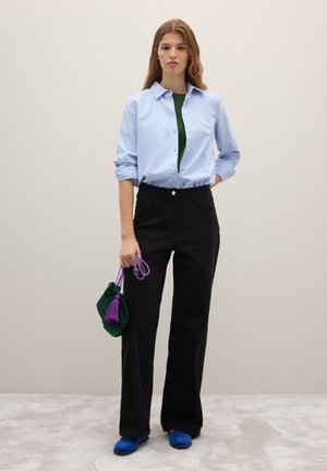 Camicia azzurro chiaro con bottoni, maglietta verde sotto, pantaloni neri a gamba larga, scarpe slip-on blu e una piccola borsa verde con nappina viola.