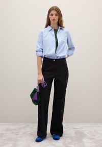Camicia azzurro chiaro con bottoni, maglietta verde sotto, pantaloni neri a gamba larga, scarpe slip-on blu e una piccola borsa verde con nappina viola.