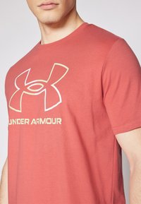 Rød bomulls t-skjorte med kort erm, med en stor hvit og gull Under Armour-logo på forsiden. Glatt tekstur, rund halsdesign.