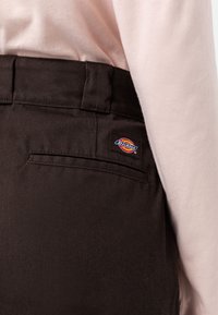 Hnědé látkové kalhoty s texturovaným povrchem, vybavené zadní kapsou a modrou, červenou a žlutou nášivkou s logem Dickies na pase.