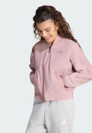 Giovane donna con capelli ricci che indossa una giacca viola chiaro con cerniera e pantaloni grigio chiaro, sorridente con le mani nelle tasche della giacca.