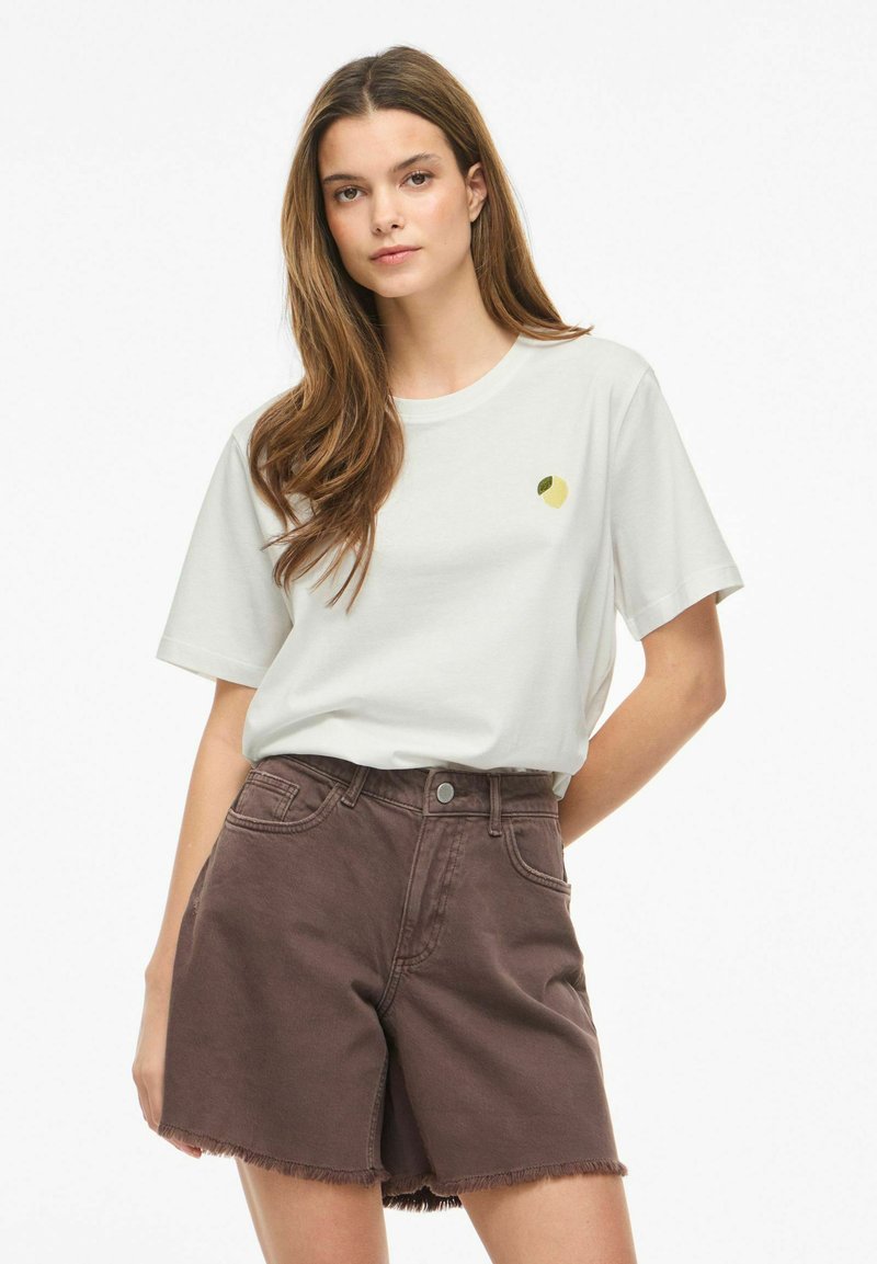 Ung kvinde med langt hår iført en hvid T-shirt med et lille gult frugtemblem og højtaljede brune denimshorts.