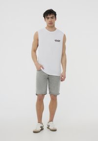 Canotta bianca con stampa grafica, abbinata a pantaloni corti grigi chiari arrotolati e sneaker bianche con accenti neri. Design semplice.