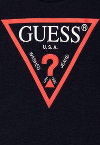 Námořnická bavlněná trička s velkým červeným trojúhelníkovým grafickým prvkem, na kterém je bílé textové logo "GUESS", slovo "WASHED" a otazník, s číslovanými detaily pod ním.