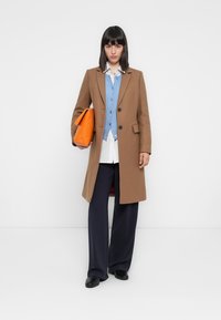 Manteau en laine marron, cardigan bleu sur une chemise blanche, pantalon large marine, chaussures noires et sac à main orange. Boutons simples et poches.