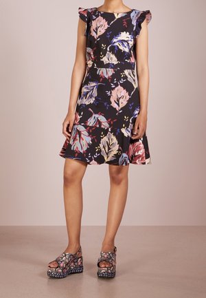 Woman wearing a black floral dress with ruffled cap sleeves and matching floral platform sandals, standing on a plain floor.