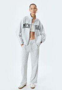 Sudadera gris con el texto "MICHIGAN", con cremallera en el frente y corte oversized, combinada con pantalones de chándal gris a juego y zapatillas blancas.
