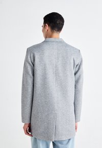 Uomo che indossa un cappotto grigio e jeans azzurri chiari, in piedi con la schiena rivolta verso la macchina fotografica su uno sfondo bianco.