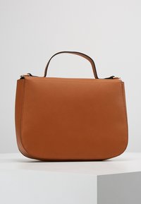 Hnědá kožená kabelka s zakřiveným dnem a krátkým uchem, vystavená na bílém podstavci na jednotném světlém šedém pozadí.