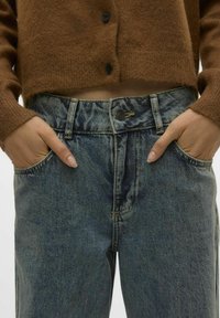 Jeans i denim med en blekt blå tvätt, hög midja och synlig sömnad. Matchas med en brun, knäppt stickad cardigan.