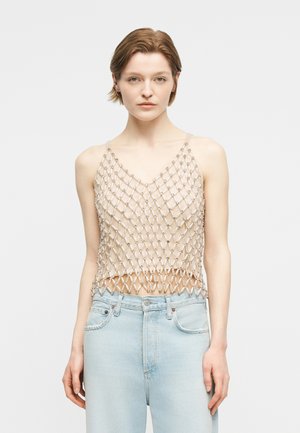 Νεαρή γυναίκα που φοράει αμάνικο μπεζ τοπ με ασημένιο δίχτυ επικάλυψης και ανοιχτόχρωμα ψηλόμεσα τζιν, στέκεται μπροστά σε απλό φόντο.