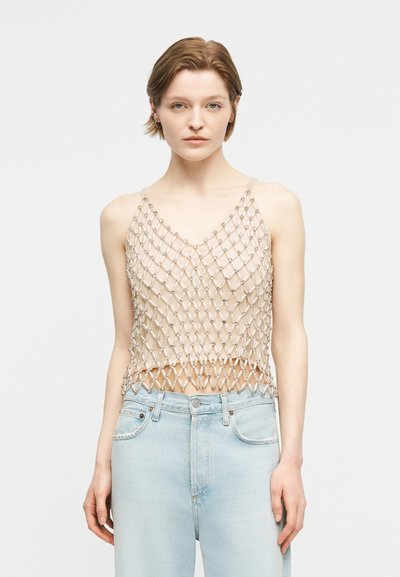 Jeune femme portant un haut beige sans manches avec une superposition en maille argentée et un jean taille haute bleu clair, debout devant un fond uni.
