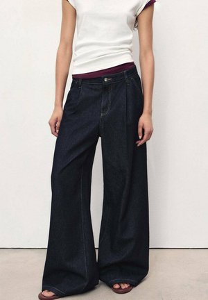 Dark blue wide-leg jeans made of cotton denim; features front pleats, a zip fly, and contrasting stitching. Worn with a white and maroon top.