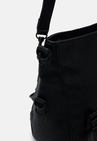 Czarna torba z teksturowanej skóry o wyraźnej, uporządkowanej formie, z podwójnymi pierścieniami do mocowania i płaskim paskiem z metalowymi ozdobami.