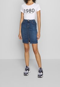 Jeanskjol i blyertsmodell med en framträdande slits, med hög midja och blå nyans. Matchas med en vit t-shirt med texten '1980 CALIFORNIA' i svart.