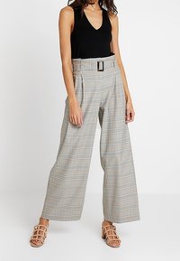 Beżowe spodnie w kratę o wysokim stanie z niebieskimi i brązowymi wzorami, z paskiem na klamrę i szerokimi nogawkami. W połączeniu z czarną bluzką i sandałami.