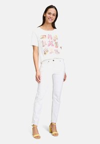 Weißes T-Shirt mit bunten Schuhillustrationen, kombiniert mit weißen Jeans und gelben hochhackigen Sandalen. Einfaches Design, lockere Passform.