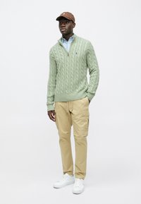 Light green cable-knit sweater with a quarter zip, paired with beige cargo pants and white sneakers. Brown cap with a logo detail.