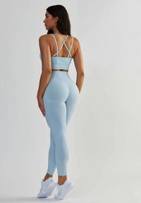 Jasnoniebieski stanik sportowy z krzyżującymi się ramiączkami, zestawiony z wysokimi legginsami w tym samym odcieniu. Gładka tekstura, dopasowany fason oraz białe sneakersy.