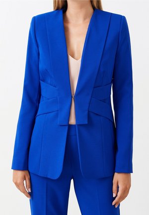Vrouw die een aansluitende kobaltblauwe blazer en bijpassende broek draagt over een lichtbeige V-hals top tegen een effen achtergrond.