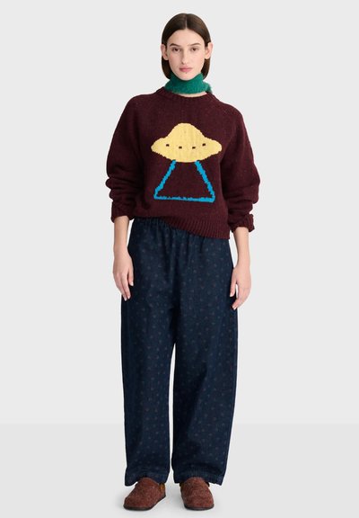 Femme portant un pull grenat avec un motif OVNI jaune et bleu, un pantalon large sombre, des chaussons marron et un chauffe-cou vert, debout devant un fond uni.