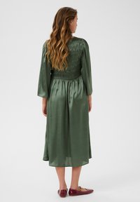 Green satin dress with a smocked back, long sleeves, and flowing skirt. Paired with burgundy flat shoes. Smooth texture, midi length.