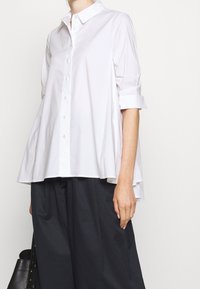 Camicia bianca con bottone, dal taglio morbido, maniche arrotolate e orlo leggermente svasato. Il tessuto appare liscio e leggermente strutturato.