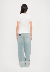 White short-sleeve t-shirt paired with light blue baggy jeans featuring a patterned patch on the back pocket, shown from the back.