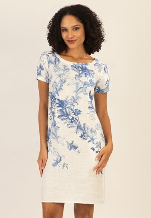 Frau mit lockigem Haar, die ein weißes kurzärmeliges Kleid mit blauen botanischen Drucken trägt und vor einem schlichten beigen Hintergrund steht.