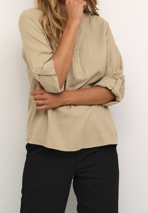 Person wearing a beige long-sleeve shirt with rolled-up sleeves and black pants, standing with one hand touching their chin.