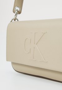 Borsa a mano in pelle beige con logo "CK" in rilievo, forma rettangolare elegante e tracolla staccabile con accessori in metallo.